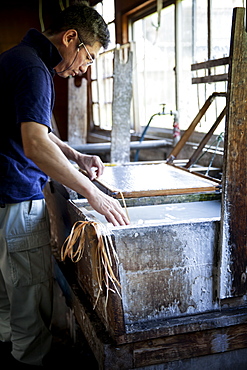 Japanese man in a workshop, holding a wooden frame with pressed pulp, making traditional Washi paper, Kyushu, Japan