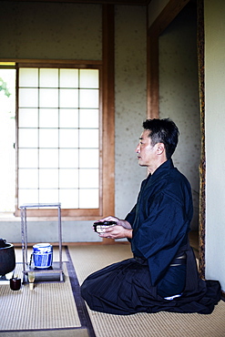 Japanese man wearing traditional kimono kneeling on floor, holding tea bowl, during tea ceremony, Kyushu, Japan