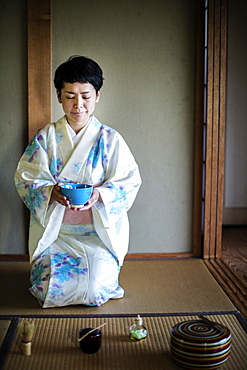 Japanese woman wearing traditional white kimono with blue floral pattern kneeling on floor during tea ceremony, holding blue tea bowl, Kyushu, Japan