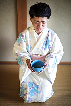 Japanese woman wearing traditional white kimono with blue floral pattern kneeling on floor during tea ceremony, holding blue tea bowl, Kyushu, Japan