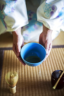 High angle close up of Japanese woman wearing traditional white kimono with blue floral pattern kneeling on floor during tea ceremony, holding blue tea bowl, Kyushu, Japan