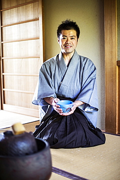 Japanese man wearing traditional kimono kneeling on floor, holding blue tea bowl during tea ceremony, Kyushu, Japan