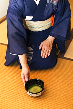 High angle view of Japanese woman wearing traditional bright blue kimono with cream coloured obi kneeling on floor, holding bowl with Matcha tea, Kyushu, Japan