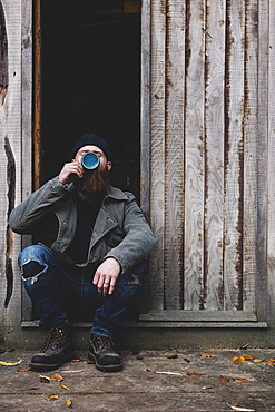 Bearded man sitting in doorway of wooden workshop, drinking tea from blue mug, Berkshire, England