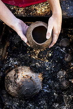 High angle close up of ceramic artist sitting next to outdoor smoke fire pit, working on vase, England