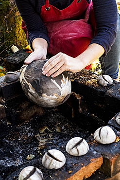 High angle close up of ceramic artist sitting next to outdoor smoke fire pit, working on vase, England