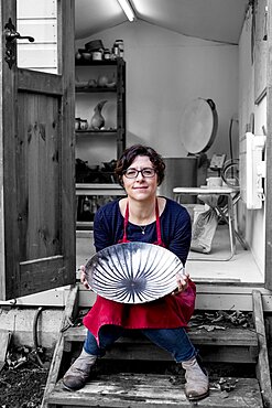 Woman wearing red apron sitting on steps outside her workshop, holding ceramic bowl with black line pattern, England