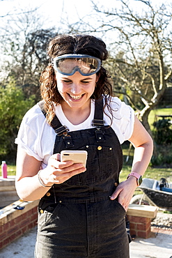 Smiling woman wearing dungarees and protective goggles checking her mobile phone, Oxfordshire, England