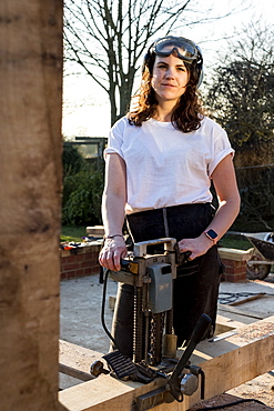 Woman wearing protective goggles holding machine, working on wooden building frame, Oxfordshire, England