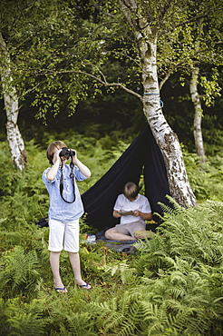 Two boys camping in New Forest. One sitting under a black canvas shelter. One boy looking through binoculars, Hampshire, England