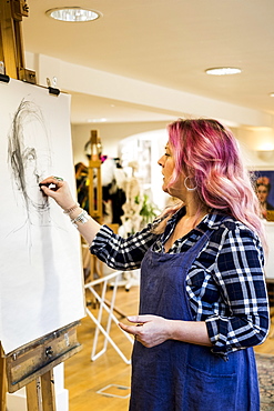 Woman with long blond wavy hair with pink streaks wearing apron standing at an easel, drawing a portrait, Oxfordshire, England