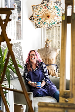 Laughing woman with long blond wavy hair with pink streaks sitting on a sofa, looking at camera, Oxfordshire, England