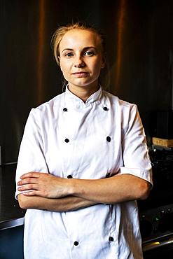 Woman wearing chef's jacket standing indoors with her arms folded, looking at camera