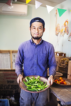 Smiling Japanese man wearing cap standing in farm shop, holding bowl with fresh okra, Kyushu, Japan