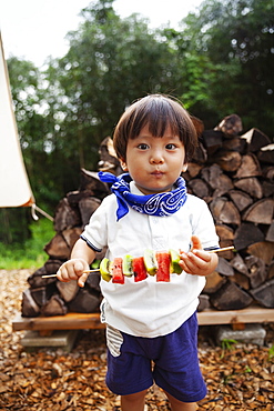 Portrait of Japanese boy holding skewer with fresh kiwi and melon, looking at camera, Kyushu, Japan