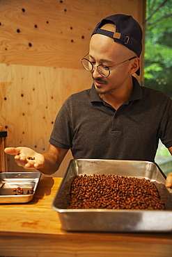 Japanese man wearing baseball cap and glasses standing in an Eco Cafe, checking freshly roasted coffee beans, Kyushu, Japan