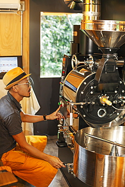 Japanese man wearing hat and glasses sitting in an Eco Cafe, operating coffee roaster machine, Kyushu, Japan