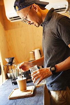 Japanese man wearing baseball cap and glasses standing in an Eco Cafe, preparing cup of coffee, Kyushu, Japan