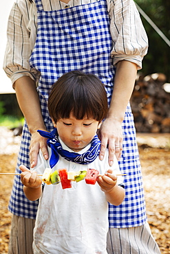 Japanese woman and boy standing outdoors, holding skewer with fresh kiwi and melon, Kyushu, Japan