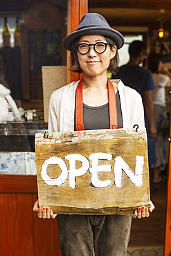 Japanese woman wearing hat and glasses standing in front of a leather shop, holding Open sign, Kyushu, Japan