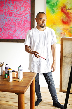 Japanese man standing in art gallery, holding paintbrush, looking at artwork on easel, Kyushu, Japan