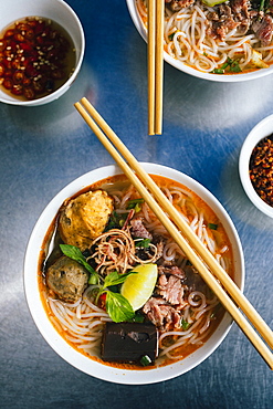 High angle close up of bowl of Bun Bo Hue with beef, crab balls, blood sausage and herbs, Vietnam