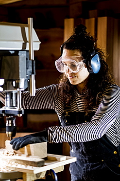 Woman with long brown hair wearing dungarees, safety glasses and ear protectors standing in wood workshop, using electric drill, Oxfordshire, United Kingdom