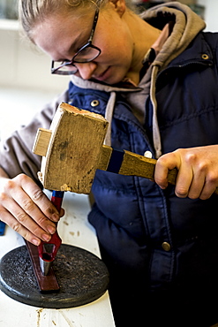 Female saddler standing in workshop, using hand tool and wooden mallet, punching holes into leather strap, Berkshire, United Kingdom