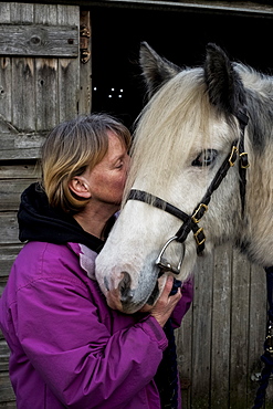 Woman standing outside stable, kissing white Cob horse, Berkshire, United Kingdom