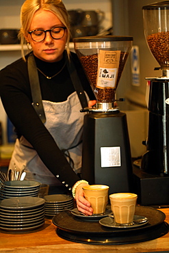 Blond woman wearing glasses and apron standing at counter in a cafe, placing two cafe lattes on a tray