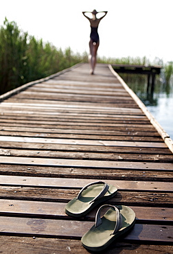 Rear view of woman wearing swimsuit walking along a jetty, hands on head, pair of flip flops in the foreground