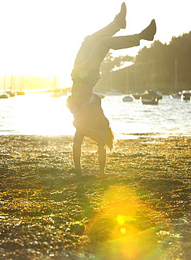 Man doing a handstand on the shore, sea and moored sailing boats in the background, sunlight, Cornwall, United Kingdom