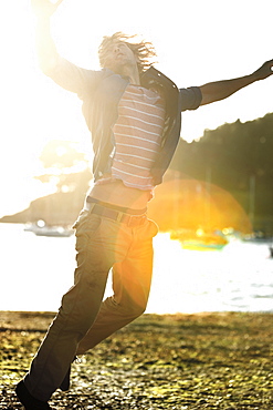 Young man jumping on a pebbly beach, arms raised, moored sailing boats in the background, sunlight, Cornwall, United Kingdom