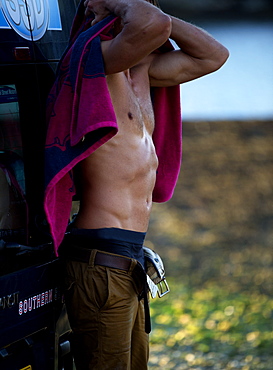 Side view of shirtless man leaning against a van, drying himself with pink towel, Cornwall, United Kingdom