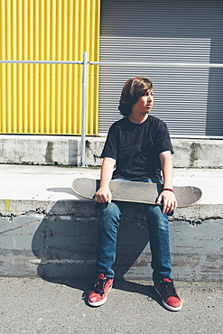 Teenage boy posing siting with skateboard in front urban warehouse