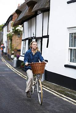 Young blond woman cycling down a village street, Oxfordshire, England, United Kingdom