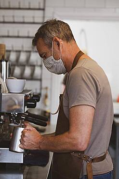 Male barista wearing brown apron and face mask working in a cafe, frothing milk