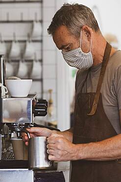 Male barista wearing brown apron and face mask working in a cafe, frothing milk