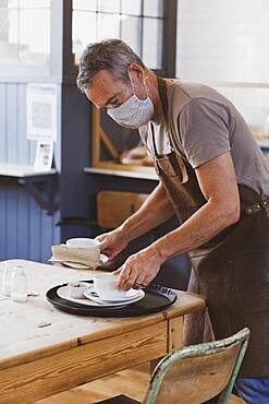 Waiter wearing brown apron and face mask working in a cafe, clearing table