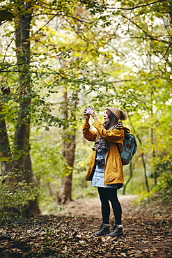 Woman standing on woodland path taking photograph using smart phone