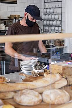 Male barista wearing black baseball cap and face mask working behind counter.