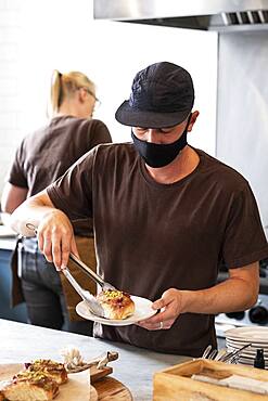 Male barista wearing black baseball cap and face mask working behind counter.