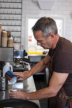 Male barista wearing brown apron, standing at espresso machine, pouring milk.