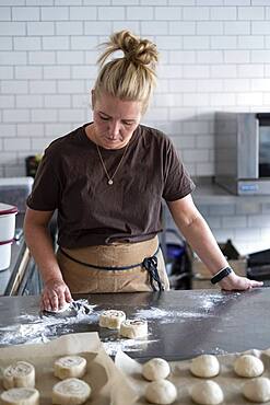 Blond woman wearing brown apron standing in a kitchen, baking danish pastries.