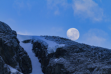 View of the nearly full moon rising over snow-covered mountains on the Antarctic Peninsula, Antarctica.