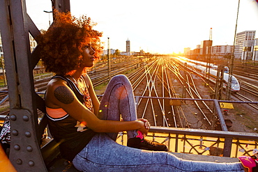 Young afro-american woman in evening light sitting on steel griders in urban scenery with tracks, Hackerbruecke Munich, Bavaria, Germany