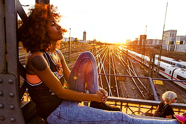 Young afro-american woman in evening light sitting on steel griders in urban scenery with tracks, Hackerbruecke Munich, Bavaria, Germany