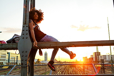 Young afro-american woman in evening light sitting on steel griders in urban scenery with tracks, Hackerbruecke Munich, Bavaria, Germany