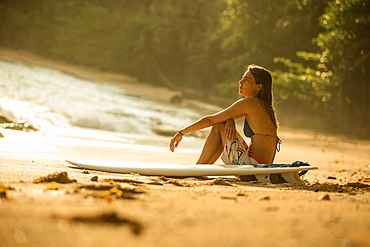 Young female surfer sitting on the beach with her surfboard, Sao Tome, Sao Tome and Principe, Africa