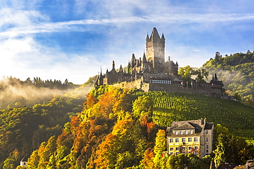 Large medieval castle on a colourful treed hillside with fog, blue sky and cloud, Cochem, Germany